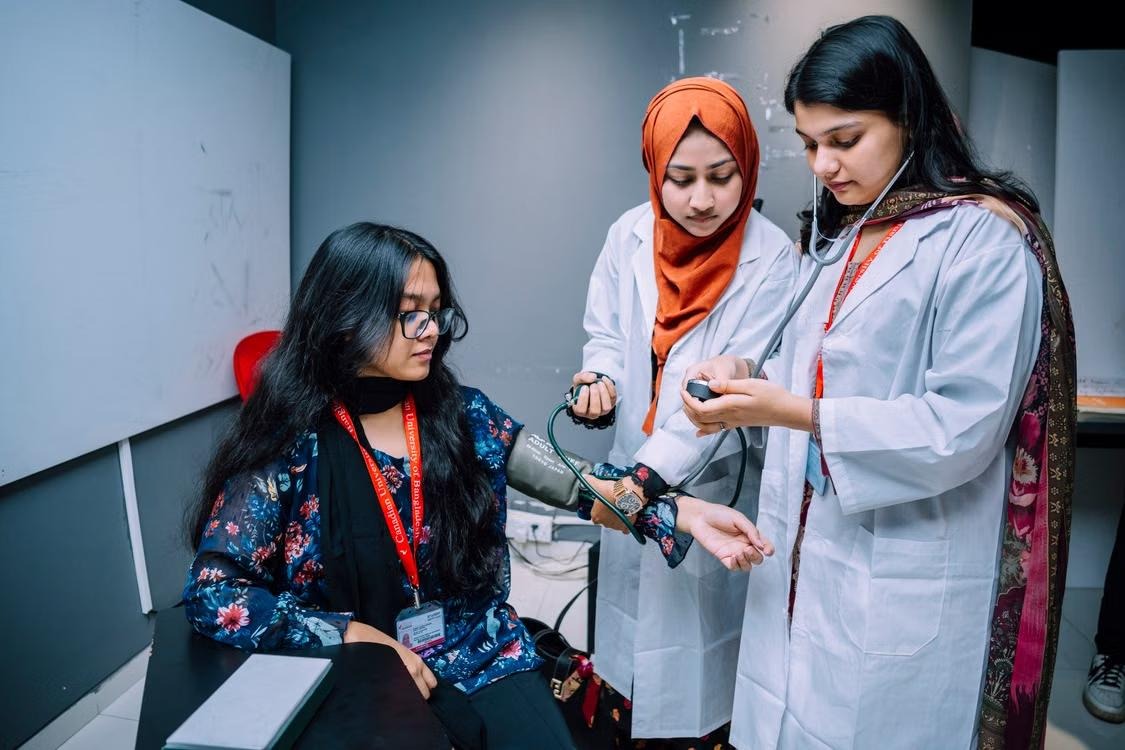 Two medical students in white coats taking a colleague's blood pressure during a clinical skills session