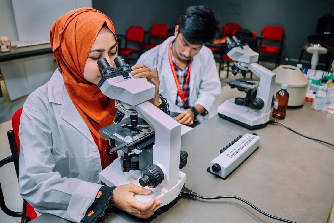 A medical student examining a slide down a microscope in a teaching laboratory, a second student at the bench behind her