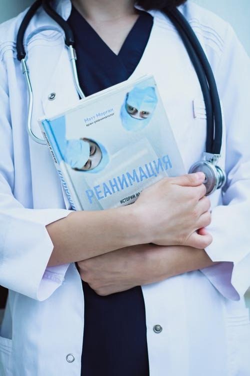 A doctor in a white coat and stethoscope holding a medical textbook against their chest