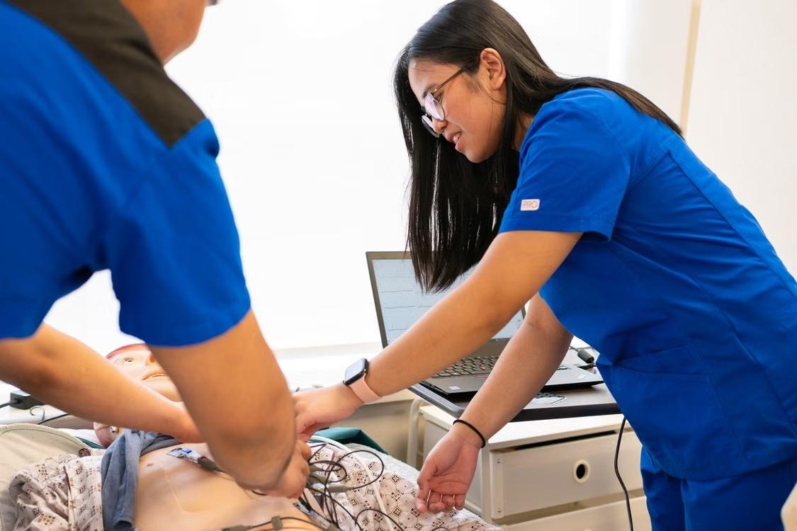 Two clinicians in scrubs practising resuscitation on a manikin, an ECG trace on the laptop beside them